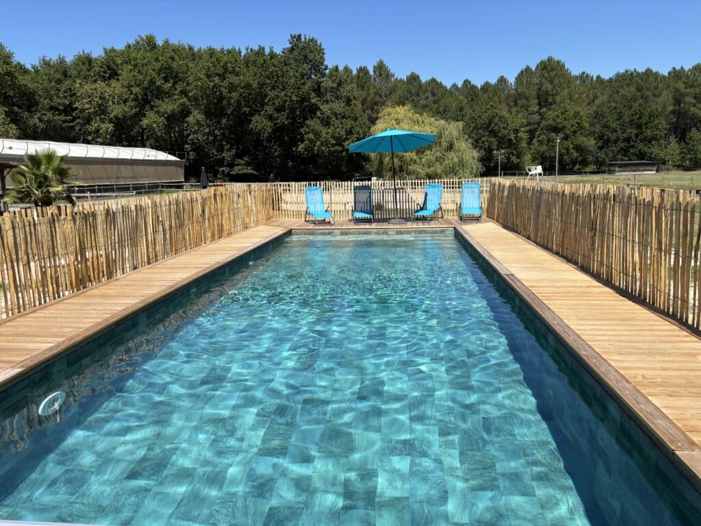 Piscine extérieure rectangulaire avec deck en bois, clôture en bambou. Quatre transats bleus, parasol turquoise, arbres luxuriants sous ciel bleu.